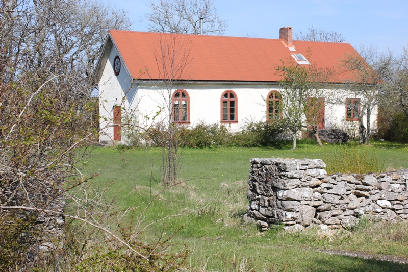 Ett vitt hus med ett rött tak, en skorsten, bruna dörrar, bruna fönster och en stenmur.