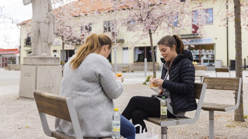Två kvinnor äter lunch utomhus på torg med blommande träd i bakgrunden