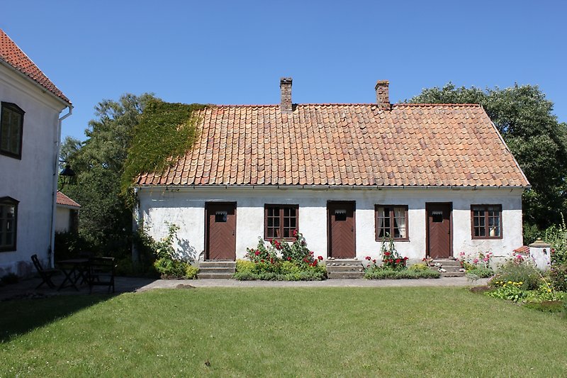 Ett vitt gårdshus med bruna fönster, bruna dörrar och ett rött tegeltak med två skorstenar.