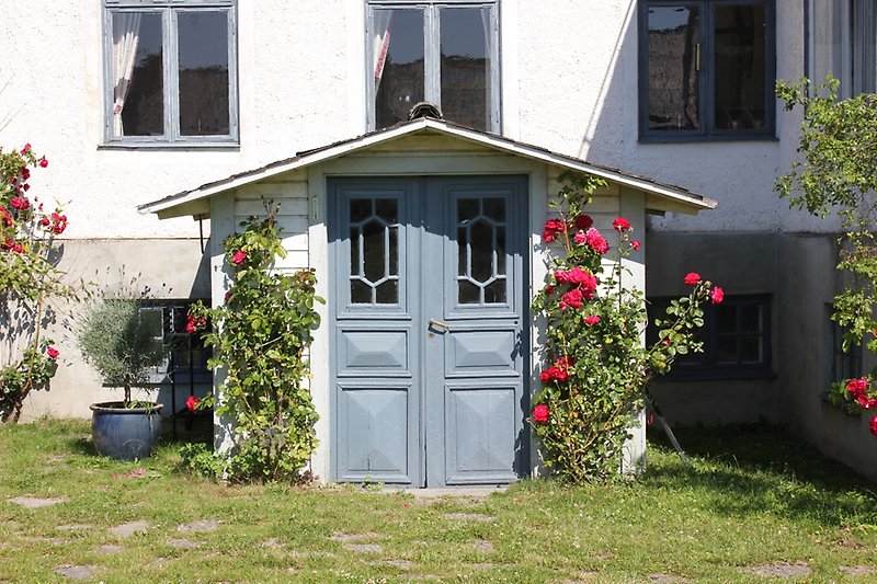 En vitt hus med ljusblåa fönster, ljusblå ytterdörr med blommande rosor på sidan.