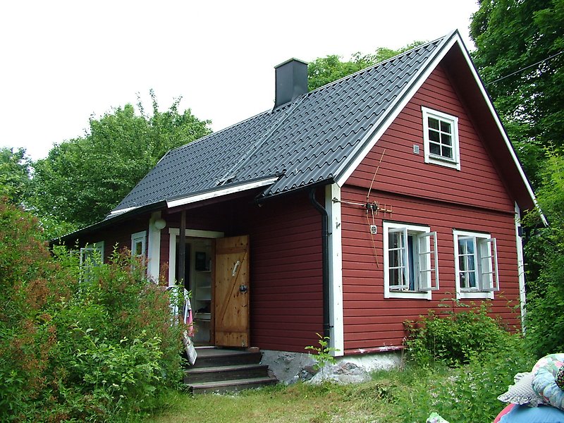 En röd stuga med vita fönster och ett svart plåttak med en skorsten.