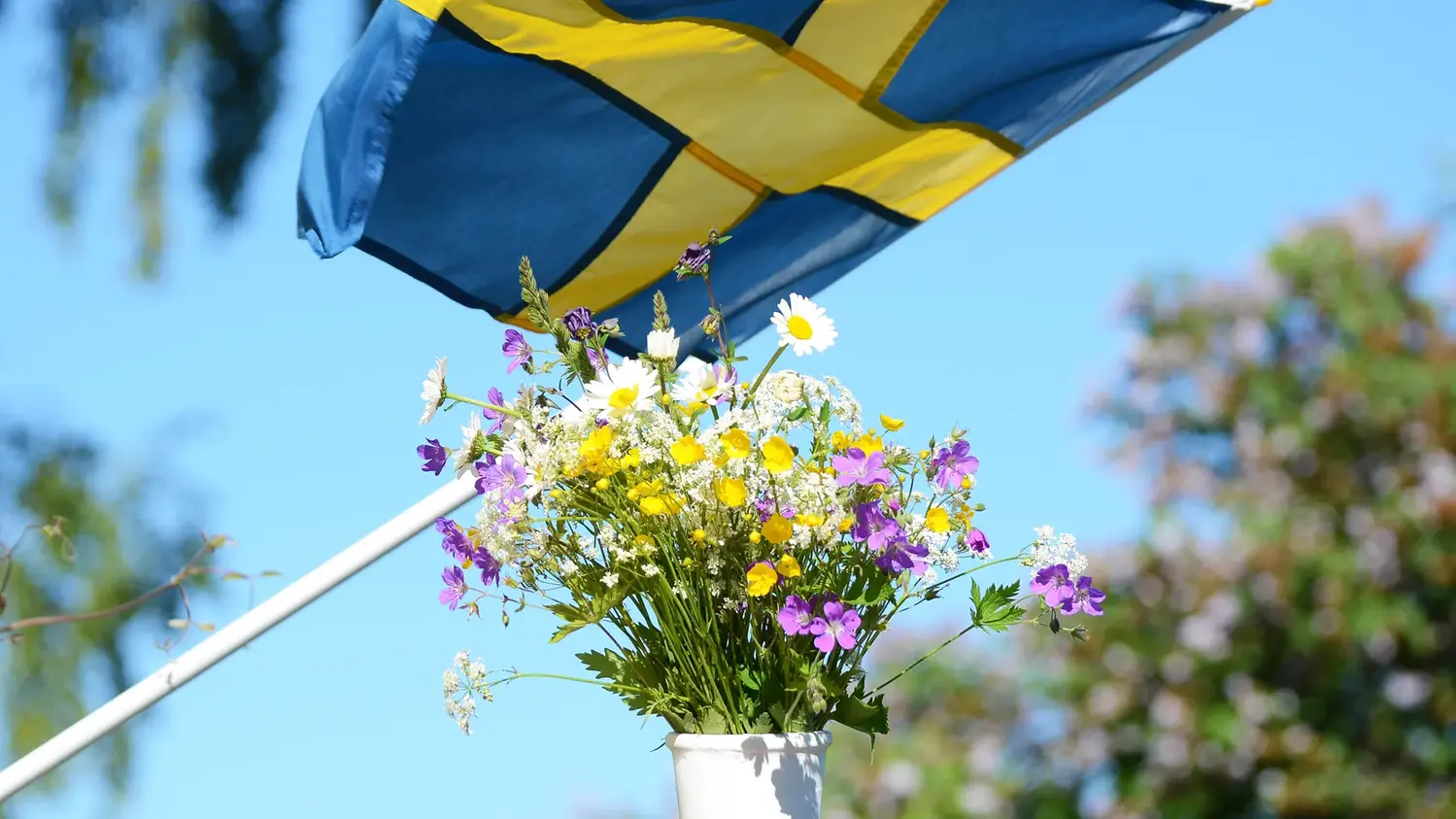 Svenska flaggan vajar i vinden mot en blå himmel och en blombukett i en vit vas.