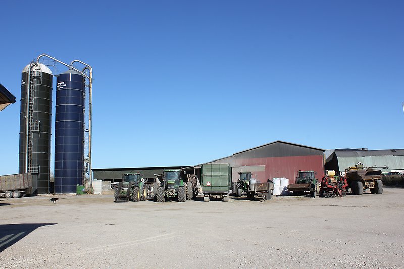 Gårdsmiljö med flera traktorer, två silos och en ladugård.