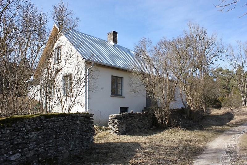På vänster sida om landsvägen står det ett vitt hus med blåa fönster, silvrig plåttak med en skorsten och med naturlig stenmur utanför byggnaden.