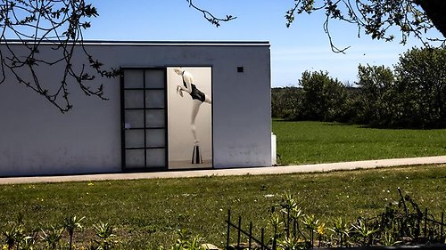 I ett grönt landskap syns ett vitt hus och genom en öppen dörr syns en staty föreställande en människa som står på ett ben