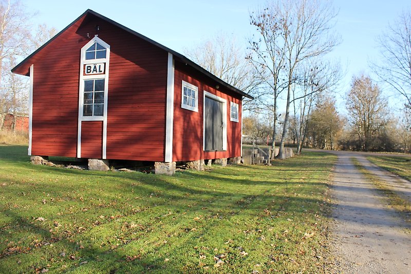 På vänster sida av grusvägen ligger det en röd träbyggnad med vita fönster och svarta dörrar.