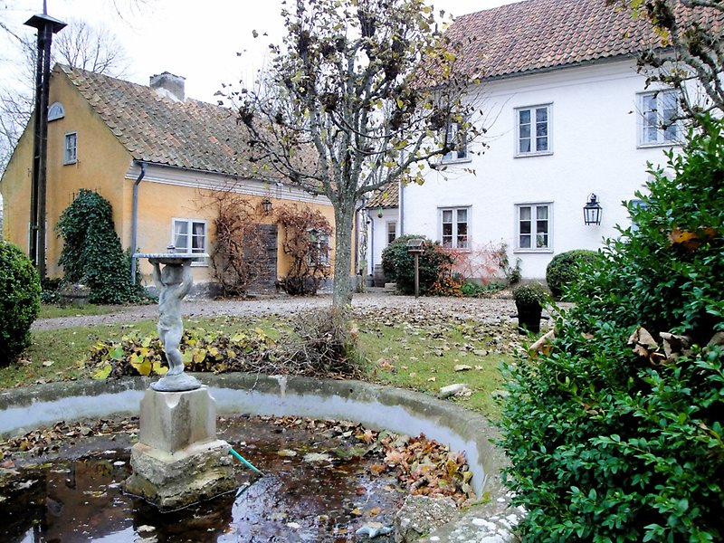 På gården finns det ett vitt tvåvåningshus, en orange stuga och en liten fontän med en skulptur.