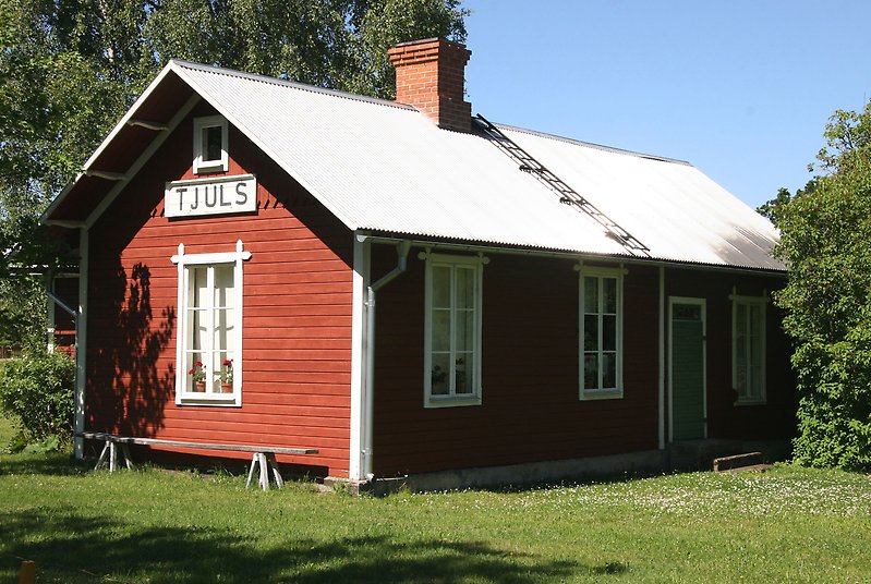 En röd stuga med vita fönster, ett plåttak med en skorsten och en skylt på gaveln med Tjuls på.
