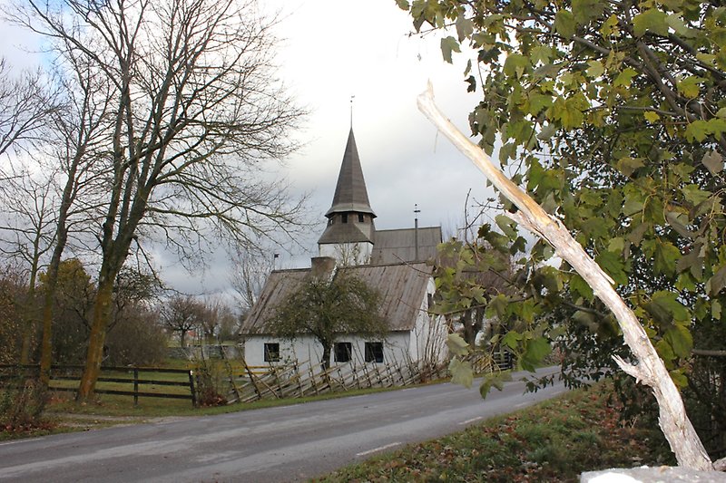 En vit kyrka med grå tak, närmare bilvägen finns det ett litet vitt hus med små fönster.