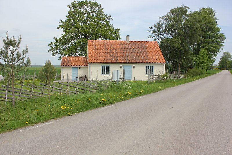 Ett beige hus med ljusblåa fönster, ljusblå dörr, ett orange tak. Huset ligger läng vägen.