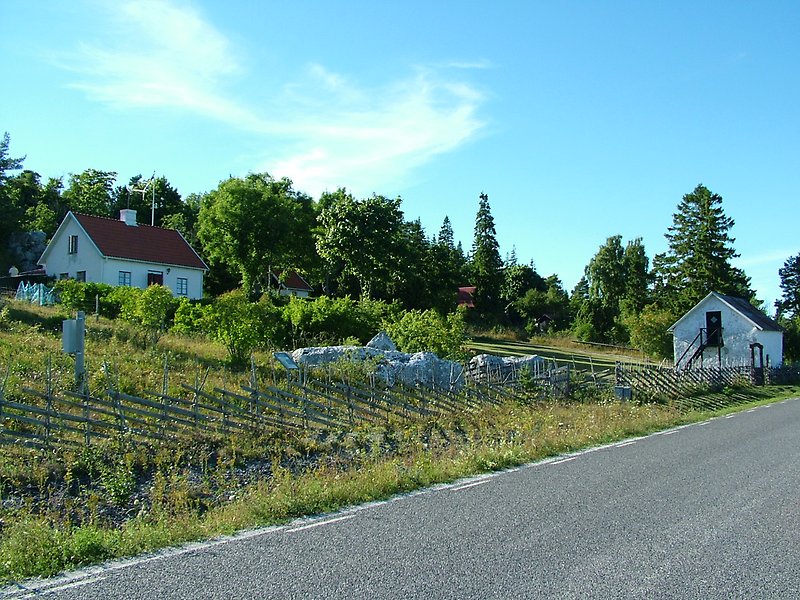 På vänster sida om bilvägen finns det ett staket och flera vita byggnader.