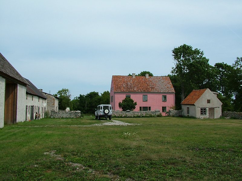 På gården finns ett rosa hus med gröna fönster, en parkerad bil, en stuga med putsade väggar och flera andra byggnader.