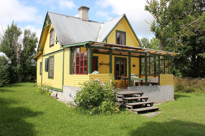 Ett sagolikt trähus i gul färg med röda fönster, gröna fönsterramar och ett plåttak.