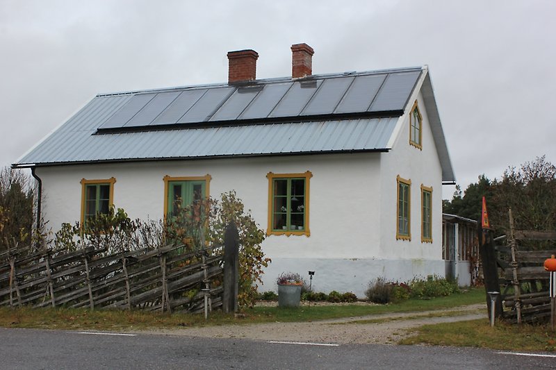 Ett litet vitt hus med gulgröna fönster, gulgrön ytterdörr, ett plåttak med solceller.