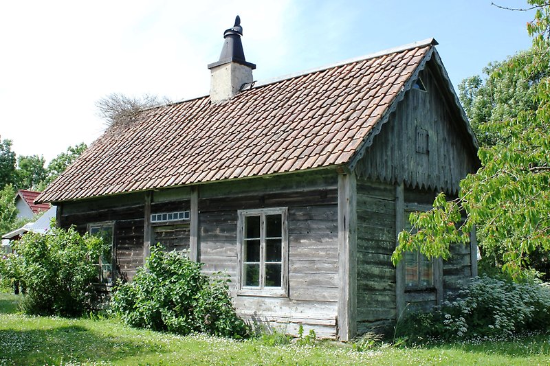 En liten stuga av obehandlad trä, vita fönster, ett rött tak och en skorsten.