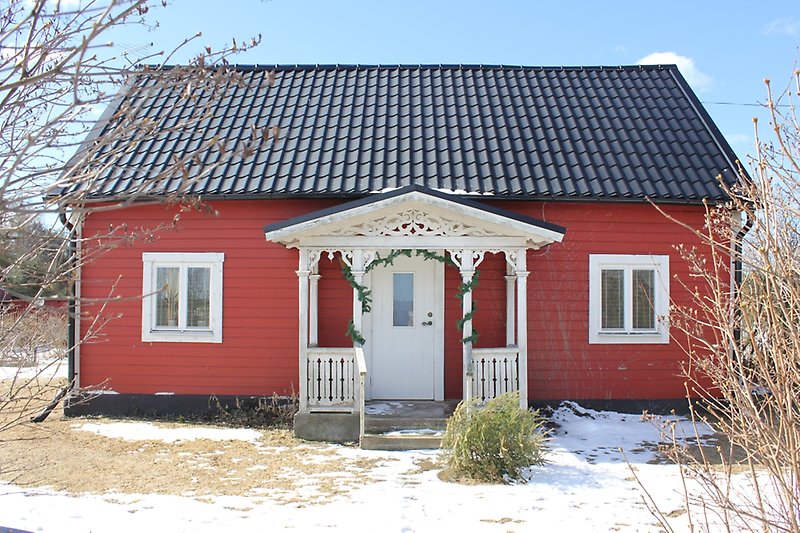 En röd stuga med en träfasad, vita fönster, vit ytterdörr och ett svart plåttak.