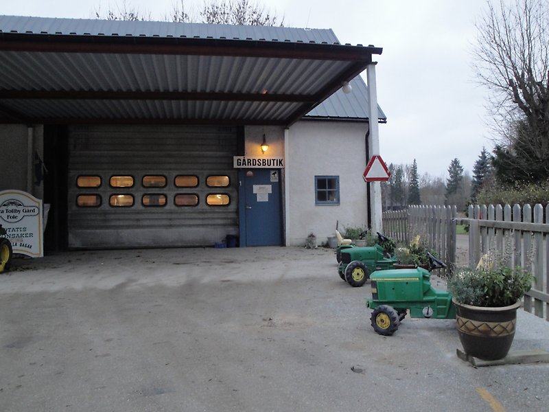 Foles gårdsbutik har blåa dörrar, blåa fönster och ett grått plåttak, utanför butiken finns det två gröna leksaks traktorer.