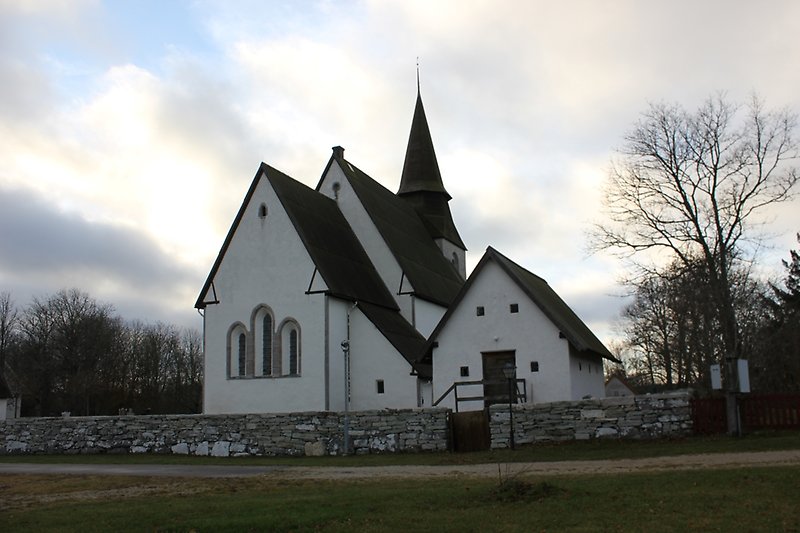 En vit kyrka med ett svart tak. Utanför kyrkan finns det en lite byggnad och en stenmur.