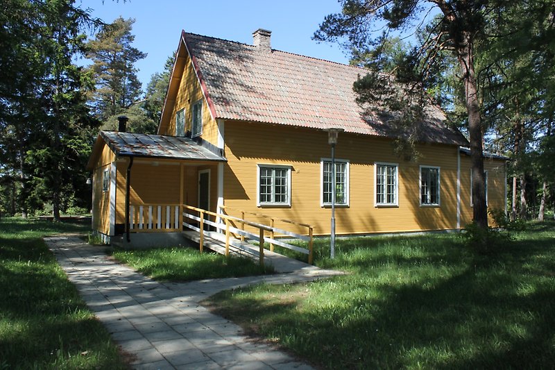I närheten av ett skogsområdet står det ett gult trähus med vita fönster och ett rött tegeltak med en skorsten.