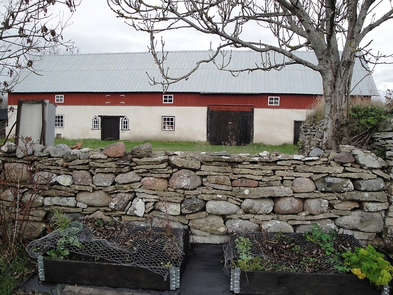 En rödvit ladugård med ett plåttak och en stenmur.